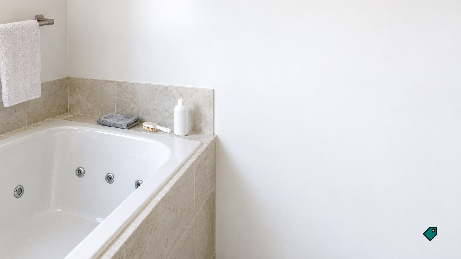 A white jetted tub sits in a beige tiled surround, with a folded gray towel, scrub brush and white pump bottle on the ledge.