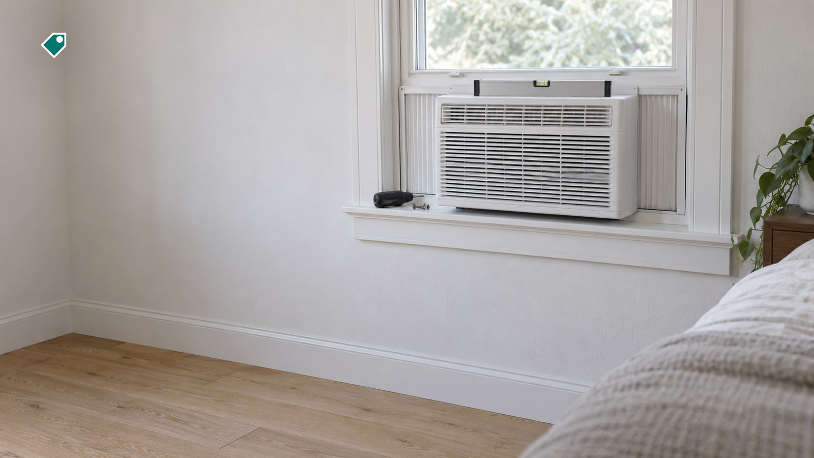 A white window air conditioner sits in a bedroom window with a level on top, and a black-handled screwdriver and a small screw rest on the sill beside it.