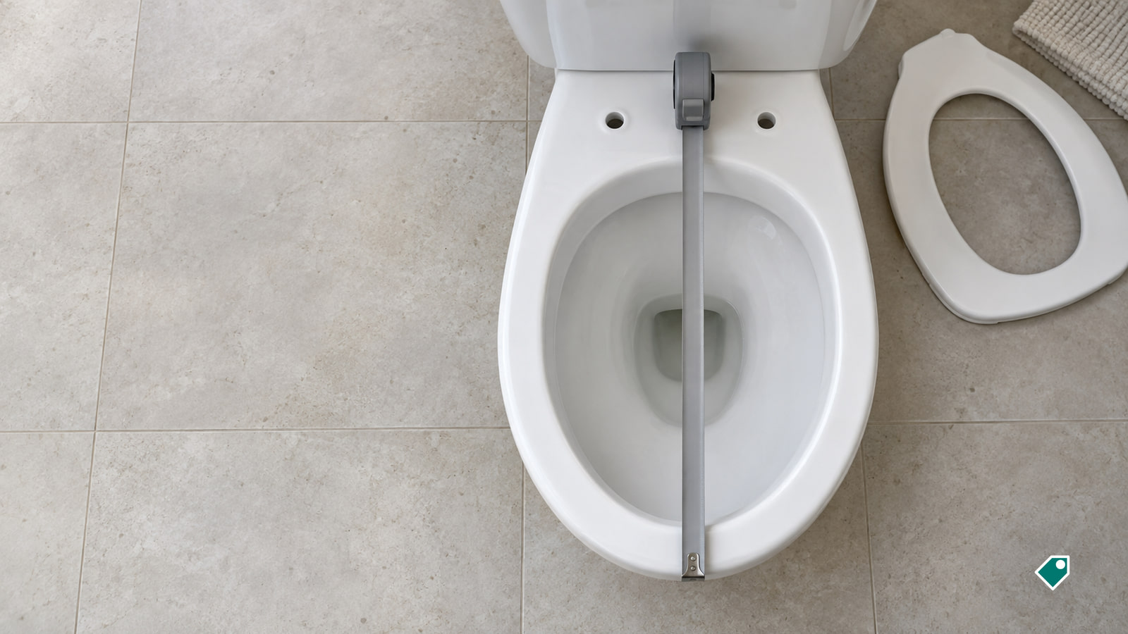 A tape measure runs from the back to the front of a white toilet bowl, with a detached seat on the tiled floor beside it.