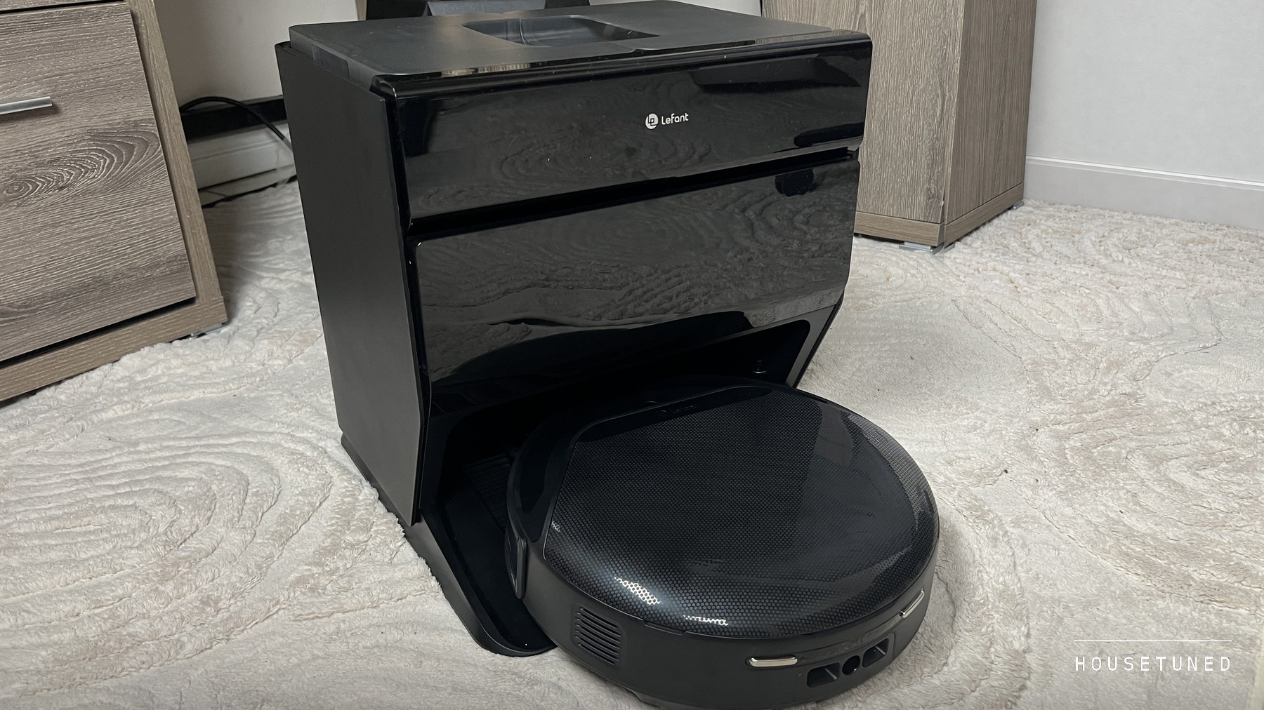The black Lefant M5 Pro robot docked in its self-emptying station on a cream shag rug, beside a light wood shelving unit