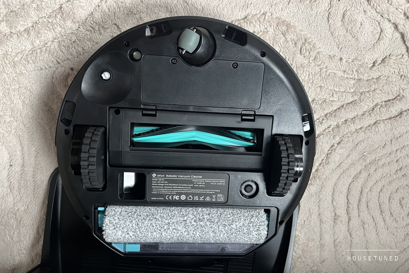 The underside of the M5 Pro on a rug: drive wheels, caster, the teal V-shaped main brush and the grey roller mop set inside the round body