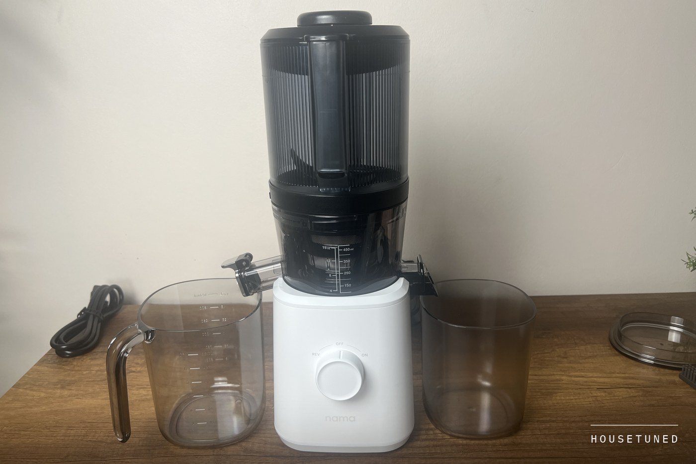 The Nama J2 assembled against a wall, the tall hopper on the white base, the juice jug and pulp container at their outlets and the cord on the counter