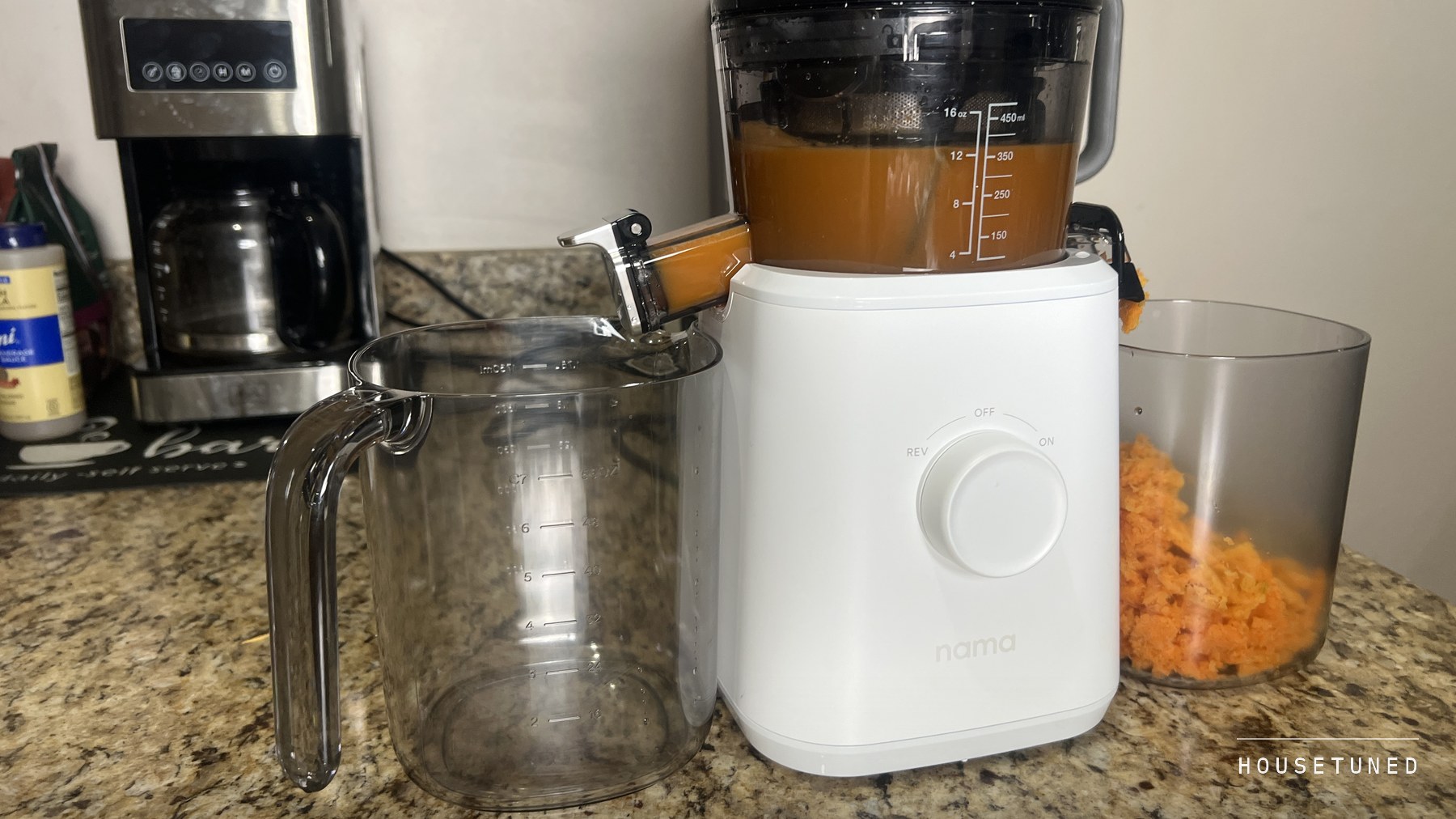 The white Nama J2 pressing on a granite kitchen counter, juice in the chamber, the jug at the juice outlet and orange pulp in the pulp container, a coffee maker behind