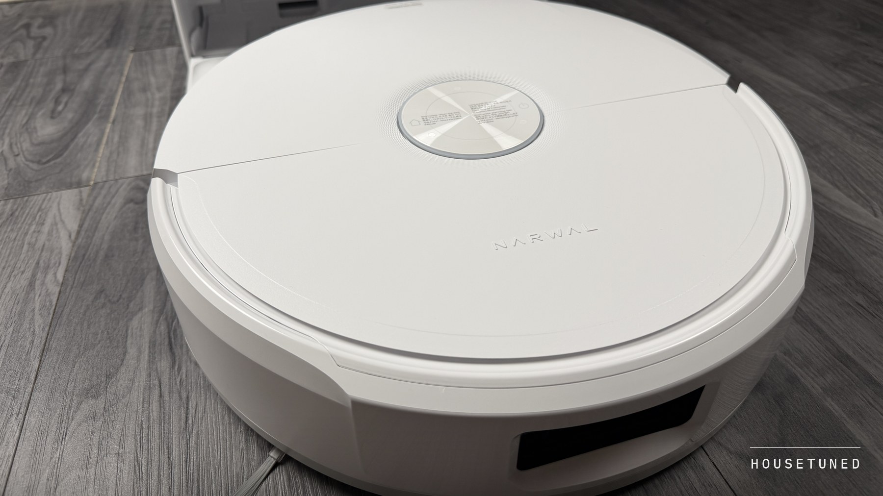 The white Narwal Freo 20 Edge robot on a grey wood-effect floor, the base of its dock just behind it