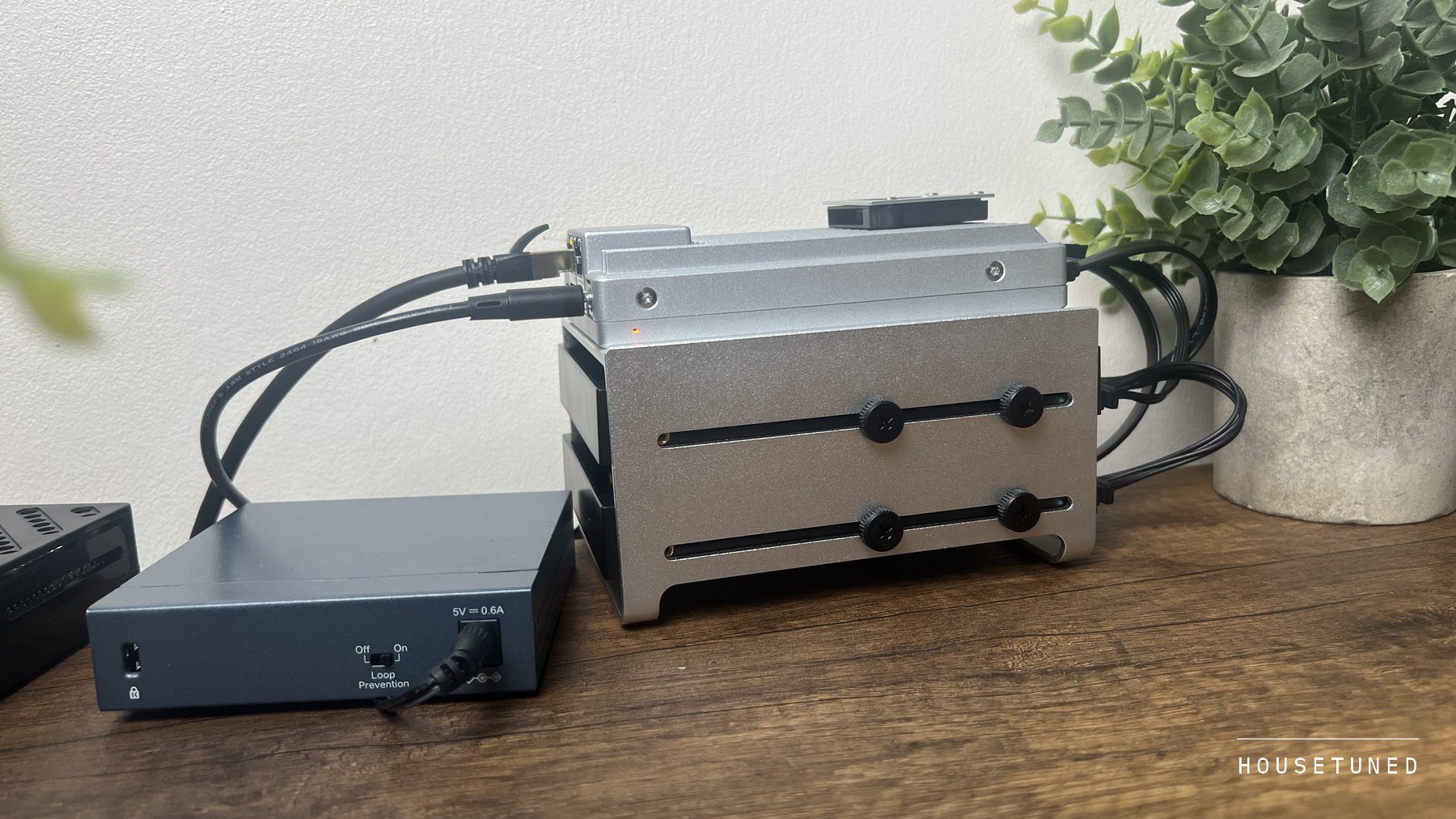 The ZimaBoard 2 in its two bay drive tray on a shelf against a white wall, a small ethernet switch beside it and a houseplant behind