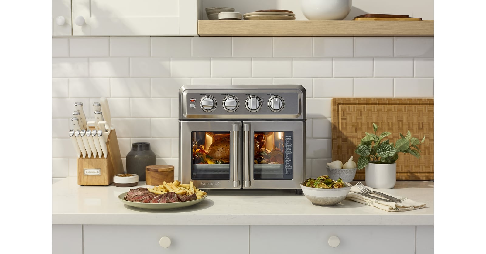 Cuisinart countertop oven with two glass doors and four control knobs sits on a kitchen counter beside prepared food.